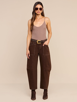 Woman wearing sunglasses, a brown tank top, the Cody Cropped Barrel-Leg Jean in high-rise dark brown non-stretch denim with a belt, and brown boots stands against a beige background.