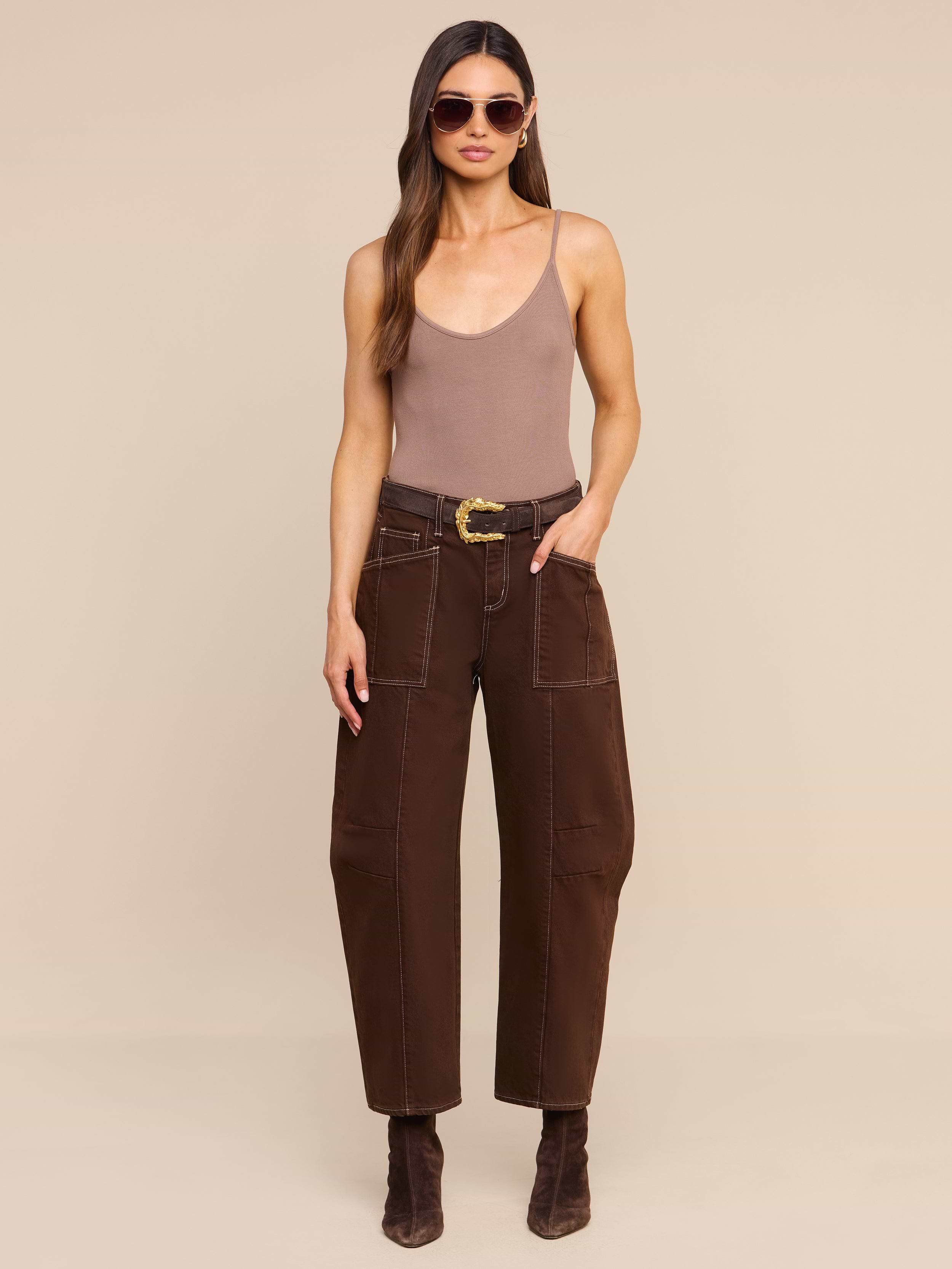 A woman stands against a plain white background wearing sunglasses, a taupe tank top, the Cody Cropped Barrel-Leg Jean in high rise dark brown with patch pockets and a belt, and dark brown boots.
