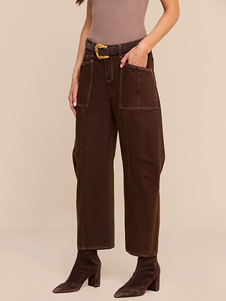A person wears the Cody Cropped Barrel-Leg Jean, a pair of high-rise brown wide-leg pants with large front pockets, styled with a black belt featuring a gold buckle, a taupe sleeveless top, and brown heeled boots. The background is beige.