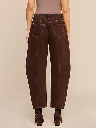 A person models the Cody Cropped Barrel-Leg Jean—a loose-fitting, high-rise brown jean with visible white stitching—paired with a brown belt and brown ankle boots. They are photographed from the back, standing against a plain beige background image shows shoulders to feet.