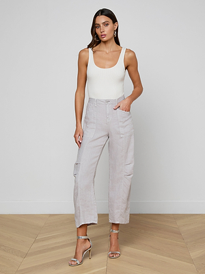 A woman with long brown hair wears a white sleeveless top and the Cody Cropped Linen Pant in light gray, featuring a high waist and pockets. She completes her look with silver heels, standing indoors against a plain white wall.