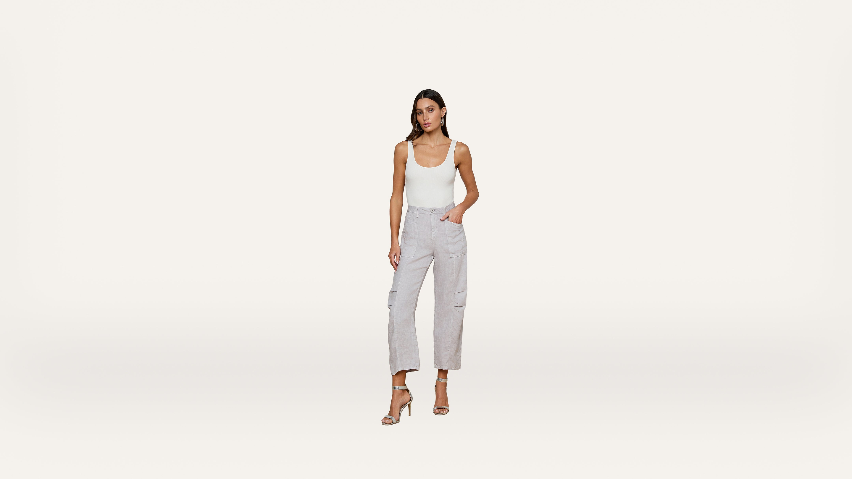 Woman standing against a white background, wearing a white tank top, light gray cargo pants, and beige high-heeled sandals, with one hand in her pocket.