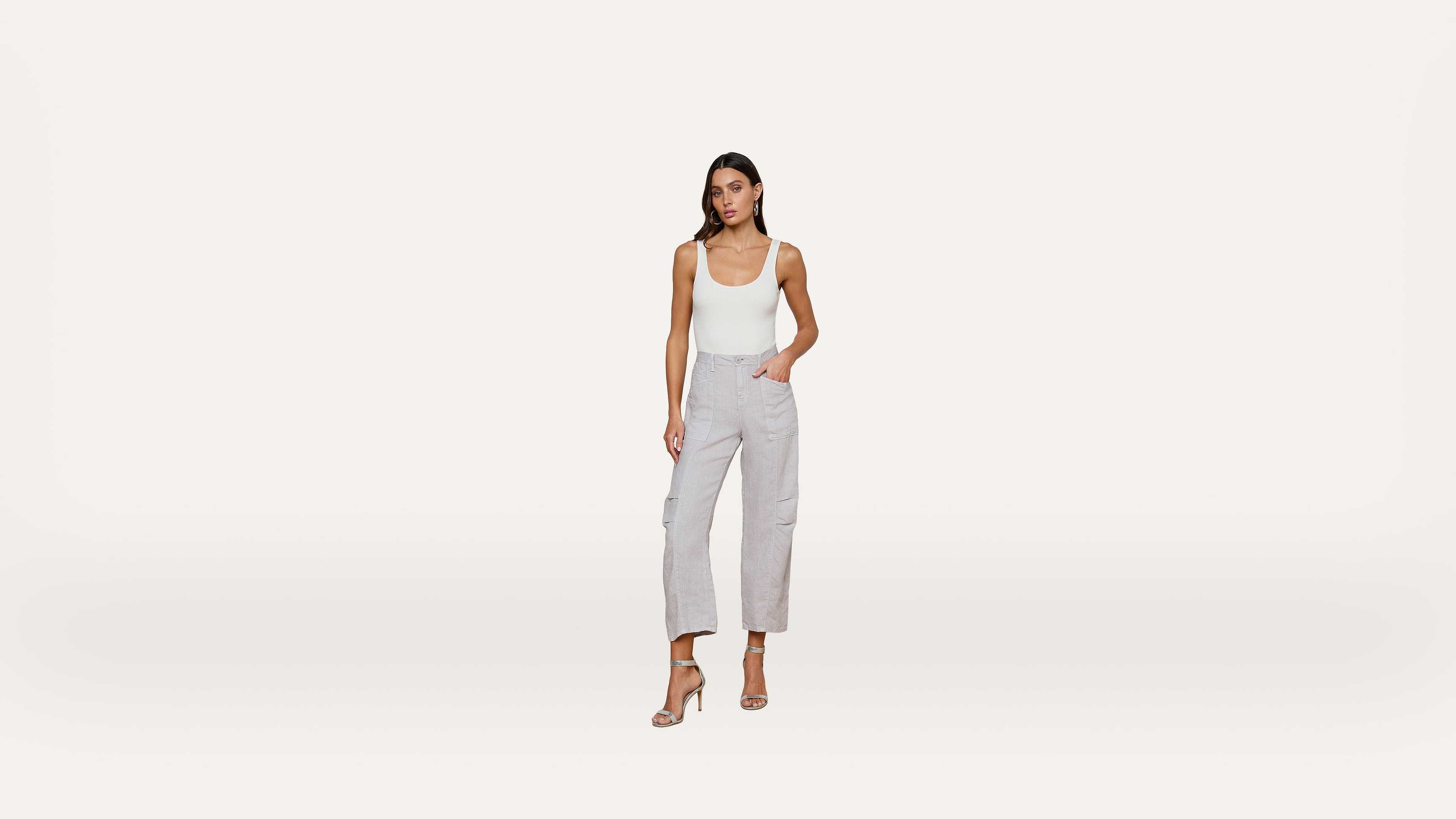 Woman standing against a white background, wearing a white tank top, light gray cargo pants, and beige high-heeled sandals, with one hand in her pocket.