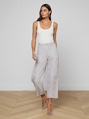 A woman with long brown hair poses indoors against a plain wall, wearing silver strappy sandals and the Cody Cropped Linen Pant in light gray—a high-waisted, barrel-leg, lightweight linen style ideal for warm weather.