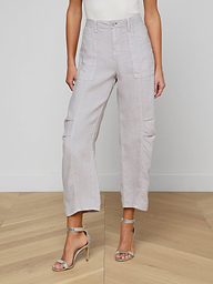 A woman stands on a wooden floor wearing the Cody Cropped Linen Pant in light gray—high-waisted with front pockets—paired with a white sleeveless top and silver high-heeled sandals. Her face is not visible.
