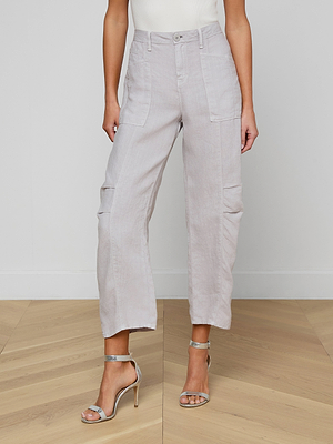 A woman stands on a wooden floor wearing the Cody Cropped Linen Pant in light gray—high-waisted with front pockets—paired with a white sleeveless top and silver high-heeled sandals. Her face is not visible.