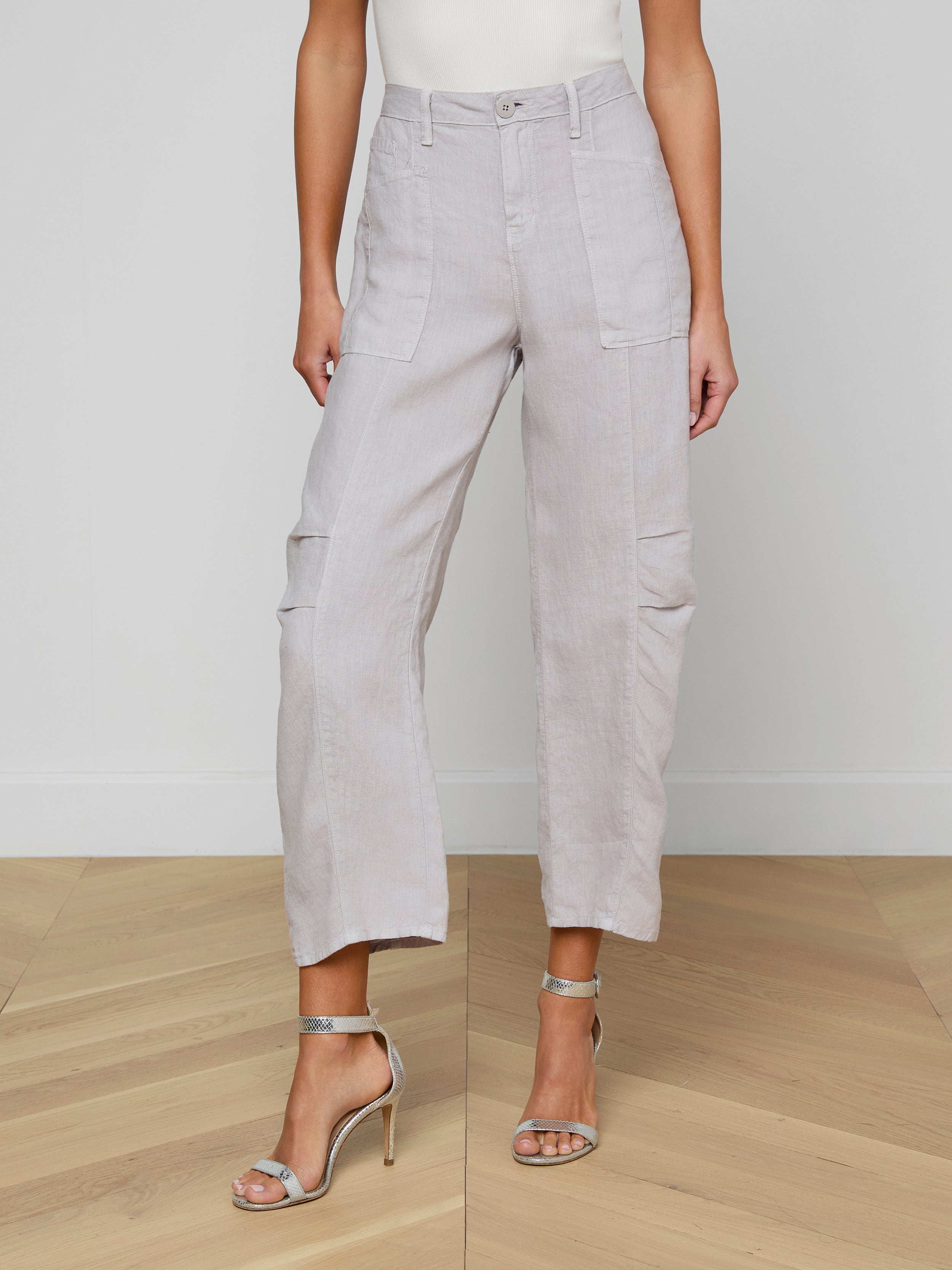 A woman stands on a wooden floor wearing the Cody Cropped Linen Pant in light gray—high-waisted with front pockets—paired with a white sleeveless top and silver high-heeled sandals. Her face is not visible.