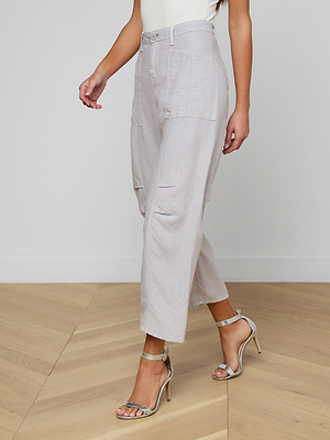 A person wears the Cody Cropped Linen Pant—light grey, high-waisted, barrel-leg trousers in lightweight linen with front pockets—paired with silver heels, standing on a light wooden floor against a plain white wall. Perfect for warm weather.