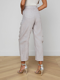 A woman stands indoors on a wooden floor, facing away from the camera, wearing the Cody Cropped Linen Pant—high-waisted, wide-leg with back pockets—paired with a sleeveless white top and silver high-heeled sandals.