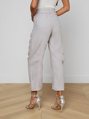 A woman stands indoors on a wooden floor, facing away from the camera, wearing the Cody Cropped Linen Pant—high-waisted, wide-leg with back pockets—paired with a sleeveless white top and silver high-heeled sandals.