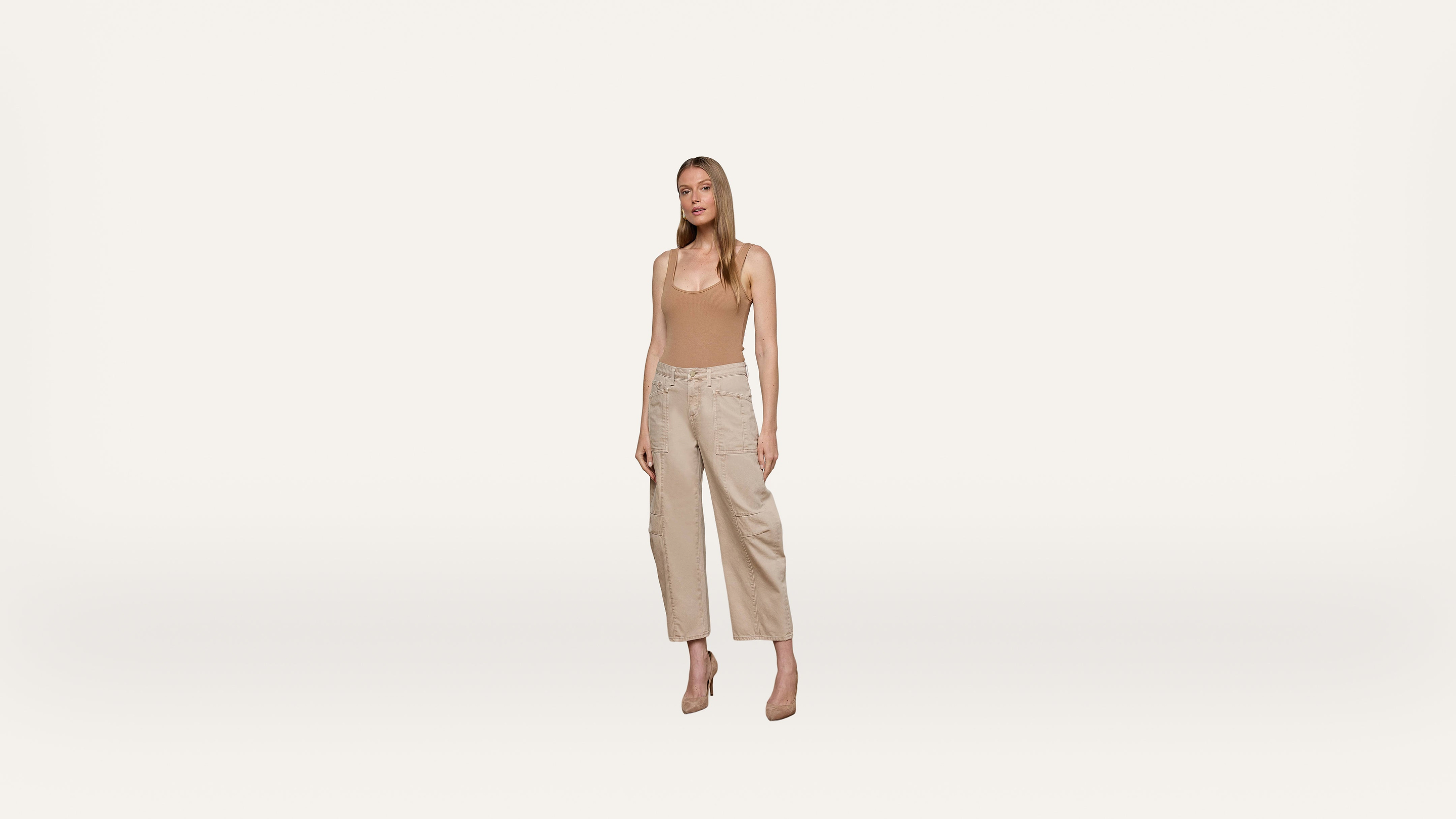 A woman stands against a plain white background, wearing a beige tank top, light-colored wide-leg pants, and beige heels. She faces forward with a neutral expression and her arms relaxed at her sides.