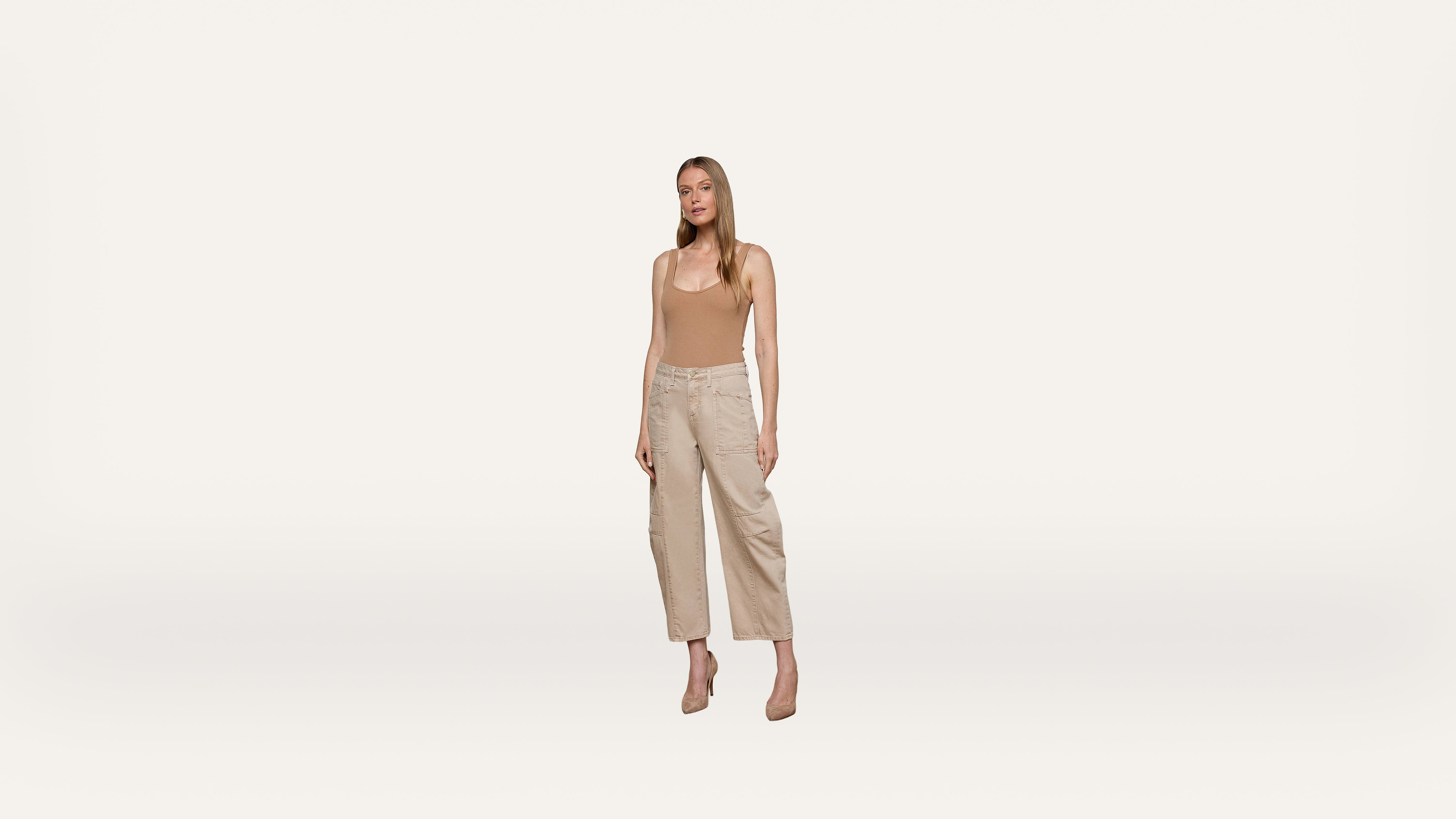 A woman stands against a plain white background, wearing a beige tank top, light-colored wide-leg pants, and beige heels. She faces forward with a neutral expression and her arms relaxed at her sides.