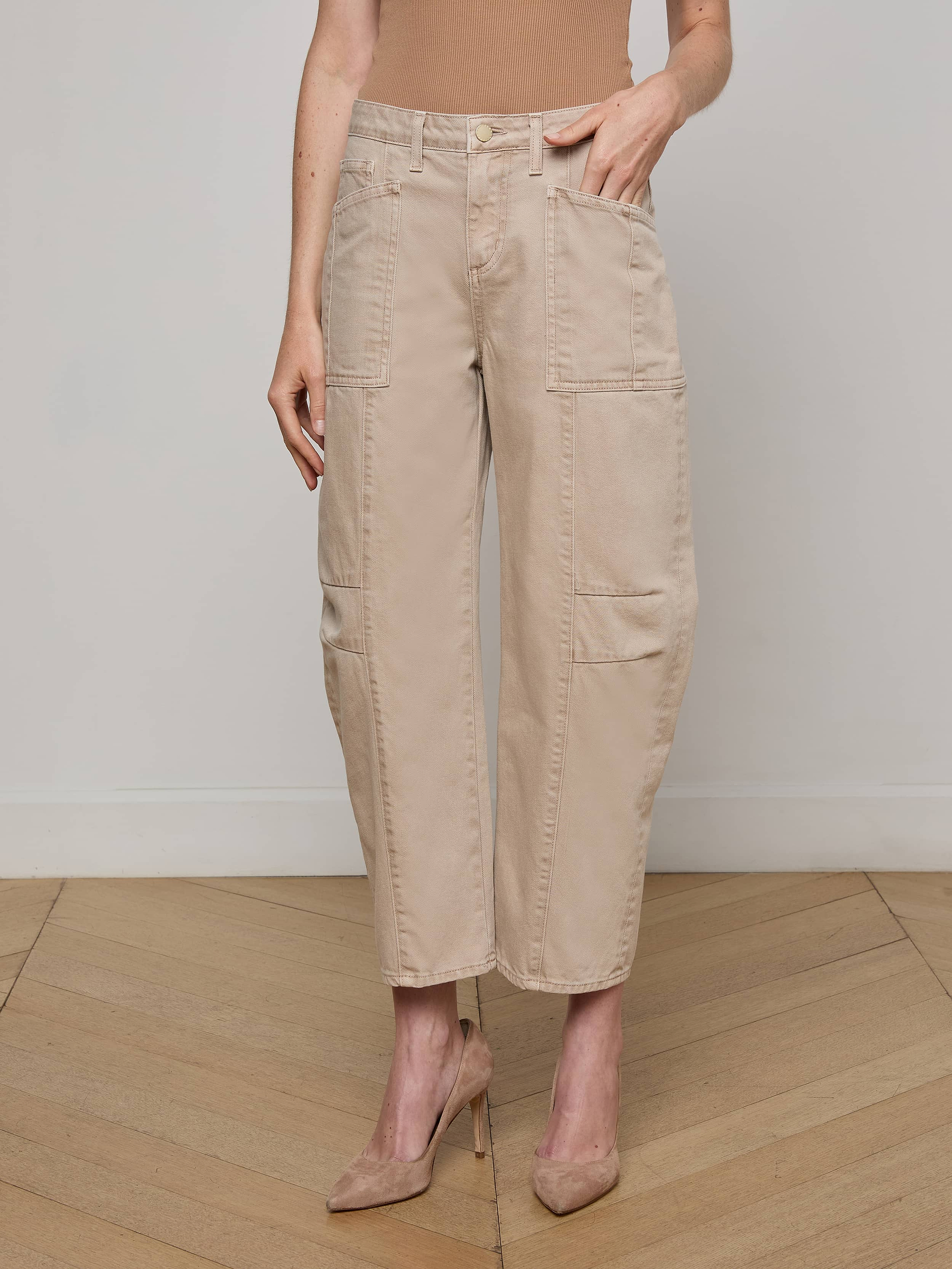 A person models the Cody Cropped Barrel-Leg Jean, featuring patch pockets and a beige hue, paired with nude heels. They stand against a plain white background, with only their lower body visible.