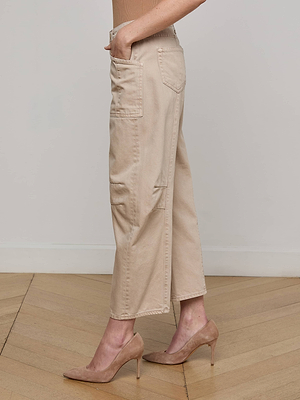 A person stands on a wooden floor wearing the L'AGENCE Cody Cropped Barrel-Leg Jean in light beige and nude high heels. The side view photo is framed from shoulder to feet, highlighting the jeans' patch pockets.