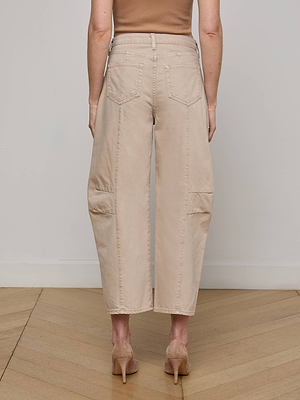 A person stands indoors on a wooden floor, shown from the back, wearing L'AGENCE Cody Cropped Barrel-Leg Jeans in light beige with patch pockets and tan high-heeled shoes.