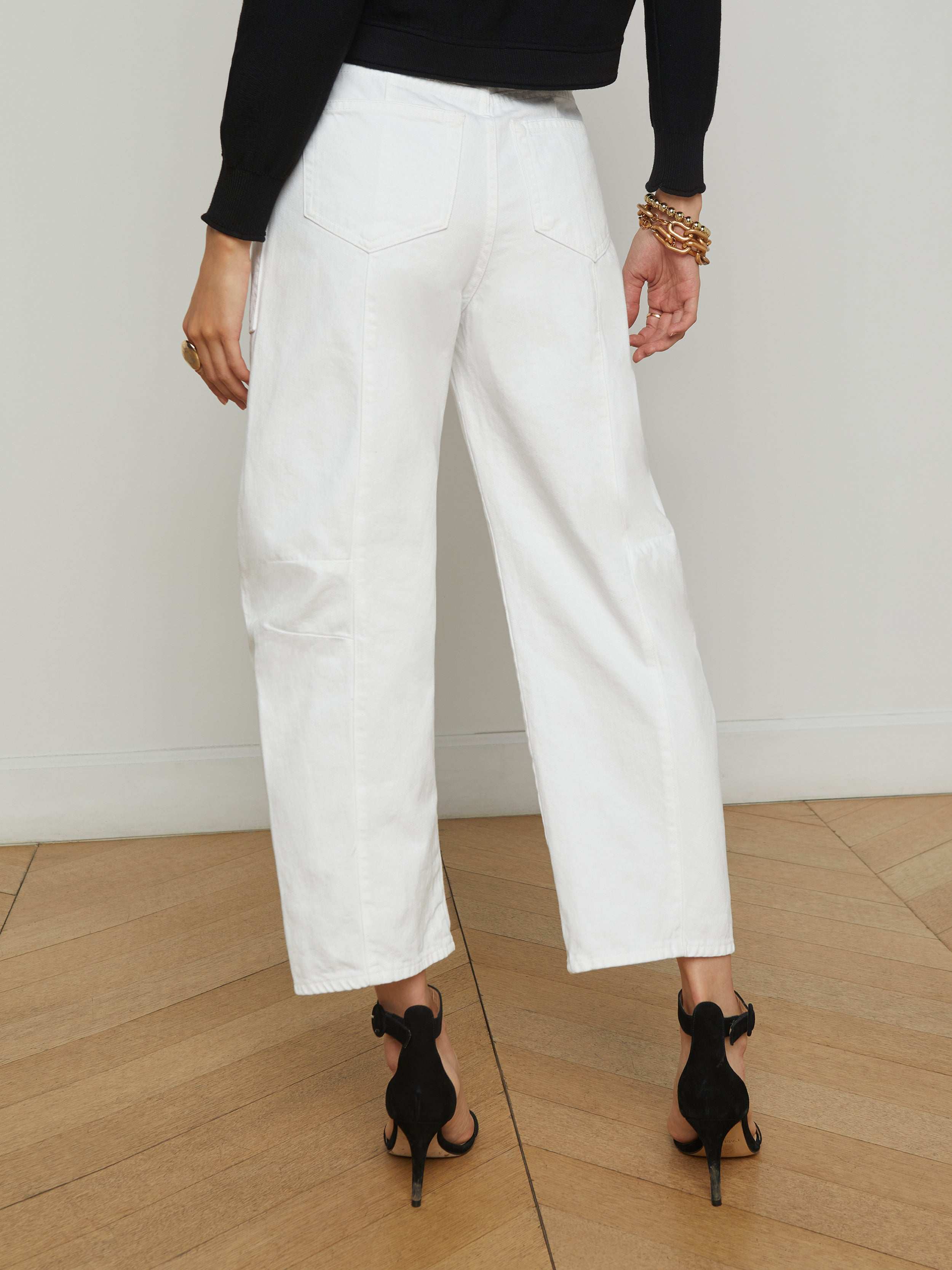 Wearing the Cody Barrel-Leg Jean in white, paired with black high-heeled sandals, a black long-sleeve top, and gold bracelets, this individual stands with their back to the camera against a plain light background.