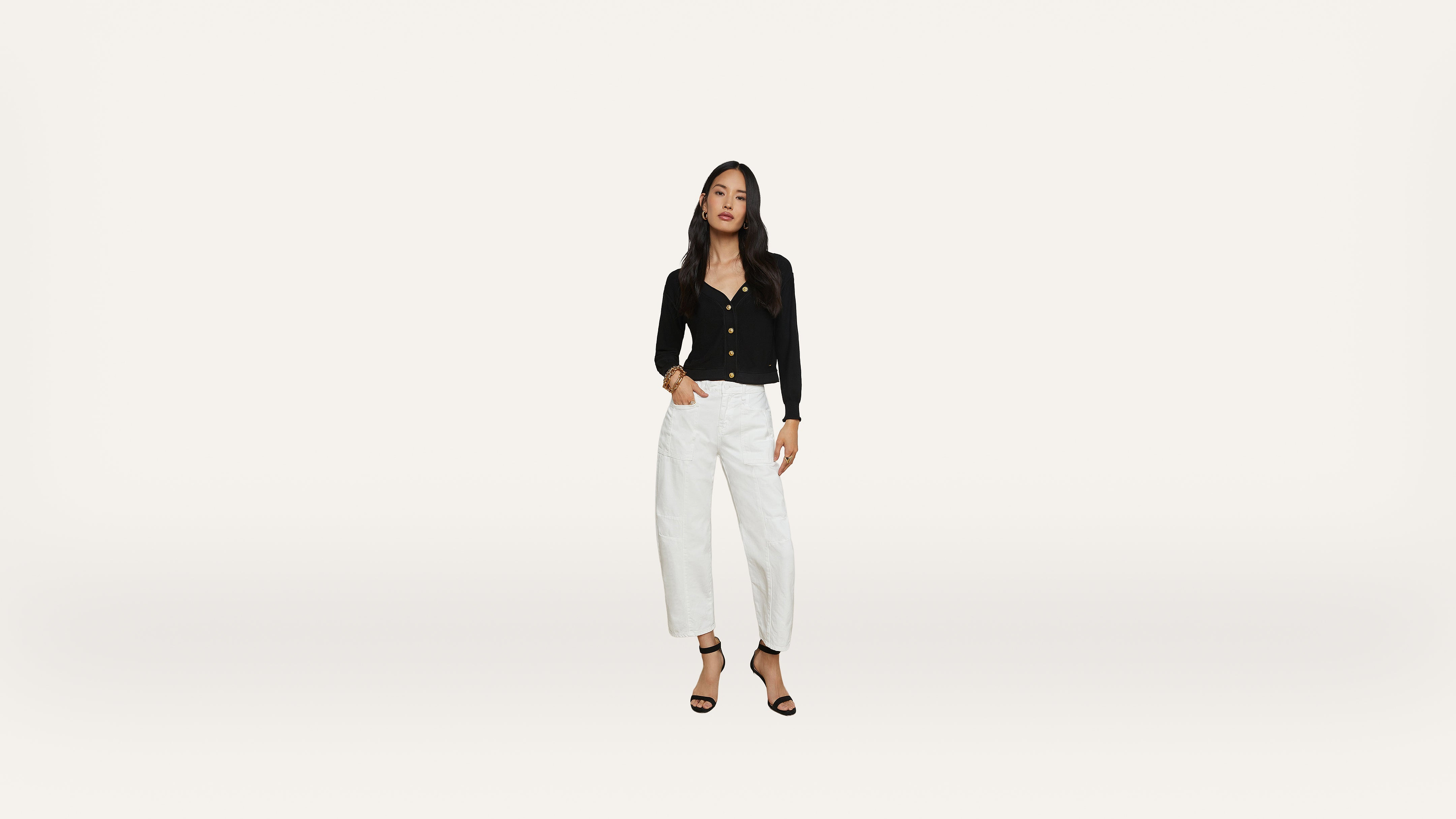 A woman stands against a plain white background, wearing a black long-sleeve top, white high-waisted pants, and black heeled sandals. She has long dark hair and a relaxed pose.