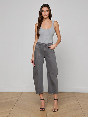 A woman with long wavy hair and a neutral expression stands indoors against a light gray wall and wooden floor, wearing a light gray tank top, Cody Cropped Barrel-Leg Jean in all-cotton denim, and black strappy heels.