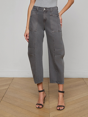 Wearing the Cody Cropped Barrel-Leg Jean in gray, paired with a gray top and black high-heeled sandals, a person stands on a wooden floor against a plain wall.