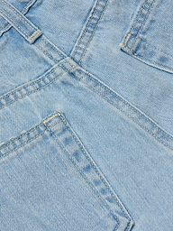 Close-up of the Teya Low-Rise Jean Short in a light blue wash, showcasing overlapping seams, pockets, and visible stitching details. The image highlights the texture, color, and structure of the denim material.