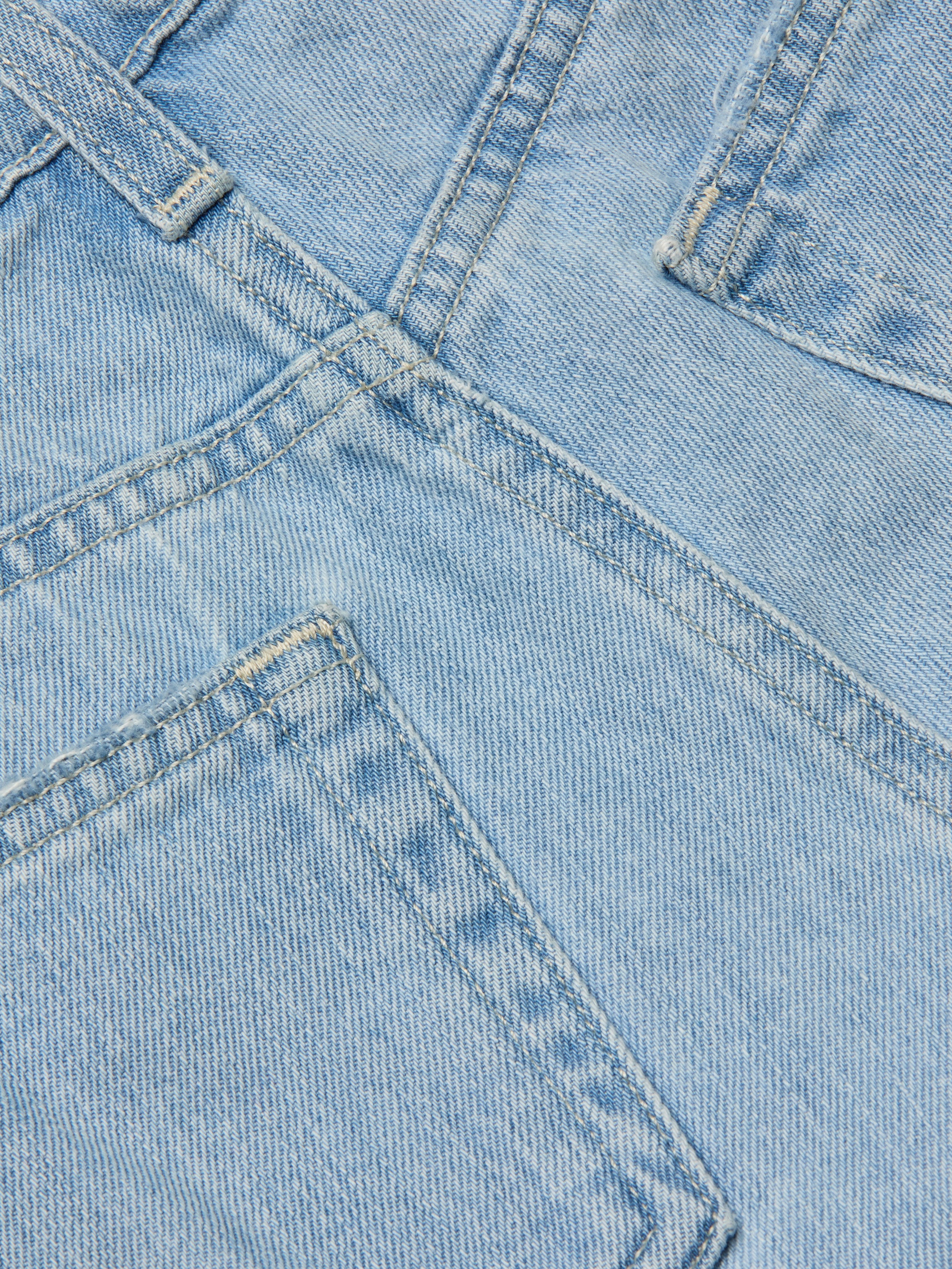 Close-up of the Teya Low-Rise Jean Short in a light blue wash, showcasing overlapping seams, pockets, and visible stitching details. The image highlights the texture, color, and structure of the denim material.