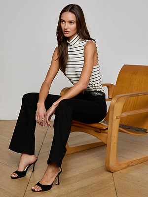 A woman with long brown hair sits on a wooden chair, wearing a sleeveless white and black striped turtleneck, black heeled sandals, and L'AGENCE Cara Low-Rise Bootcut Jeans in a minimalist room with a wooden floor.