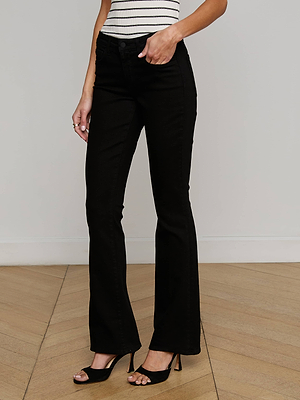 A woman wears the L'AGENCE Cara Low-Rise Bootcut Jean in black, paired with a white sleeveless striped top and black high-heeled sandals, standing on a light wood floor against a plain white wall.