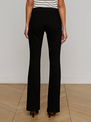 A person wearing L'AGENCE Cara Low-Rise Bootcut Jeans and black heeled shoes, photographed from behind on a wooden floor against a plain light-colored wall.