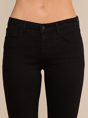 Primer plano de una persona que lleva unos vaqueros «Cara Low-Rise Bootcut» de denim elástico negro, con cinturilla, bolsillos delanteros y cierre de botón. Se ve la parte media del abdomen sobre un fondo beige liso.