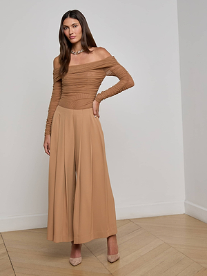 A woman stands indoors in the L'AGENCE Novalynn Cropped Pleated Pant, styled with a tan off-the-shoulder long-sleeve top, nude heels, and a necklace against a plain white wall and light wood floor.
