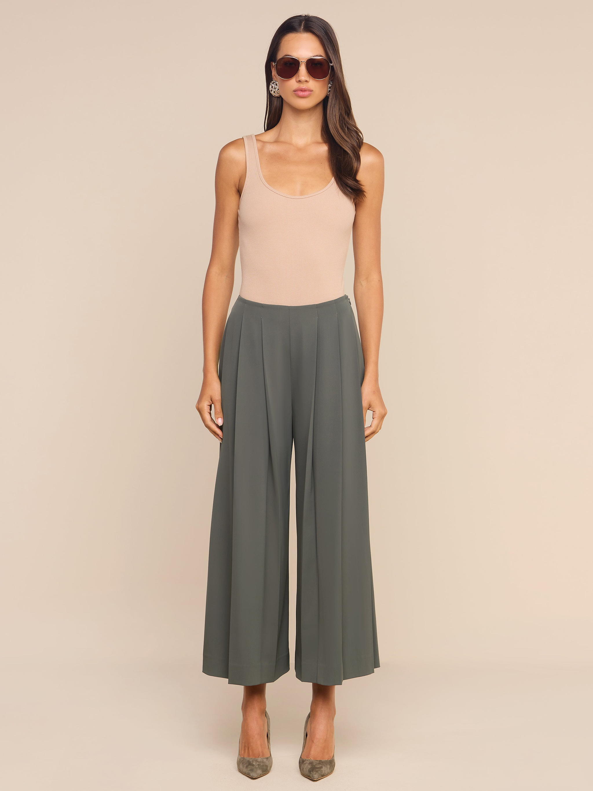 Woman wearing sunglasses, a beige sleeveless top, and high-rise, wide-leg gray Novalynn Cropped Pleated Pant stands facing forward against a plain white background. She has long brown hair and is wearing pointed-toe heels.