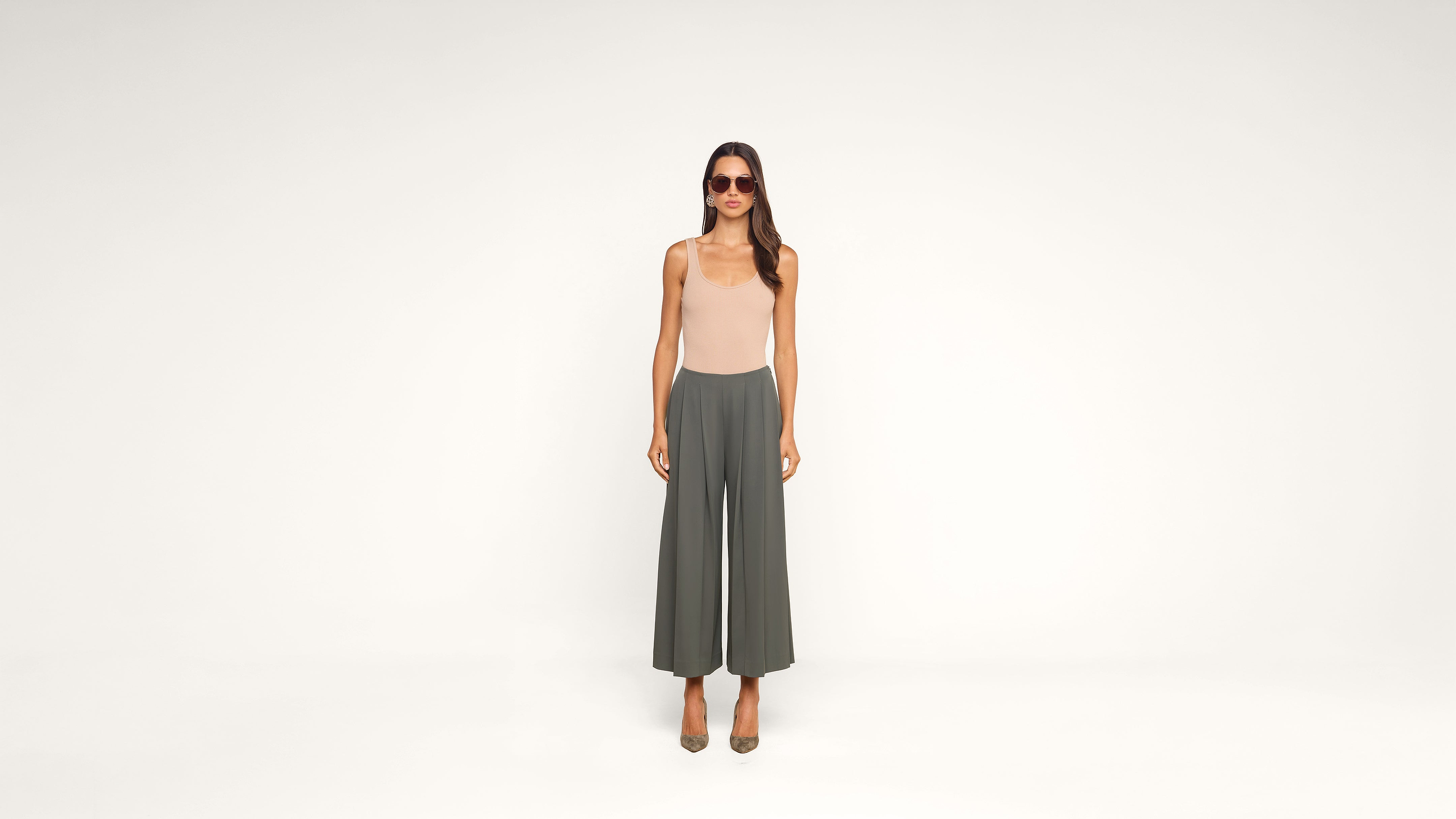 Woman in sunglasses, beige sleeveless top, and wide-leg gray pants stands facing forward against a plain white background.