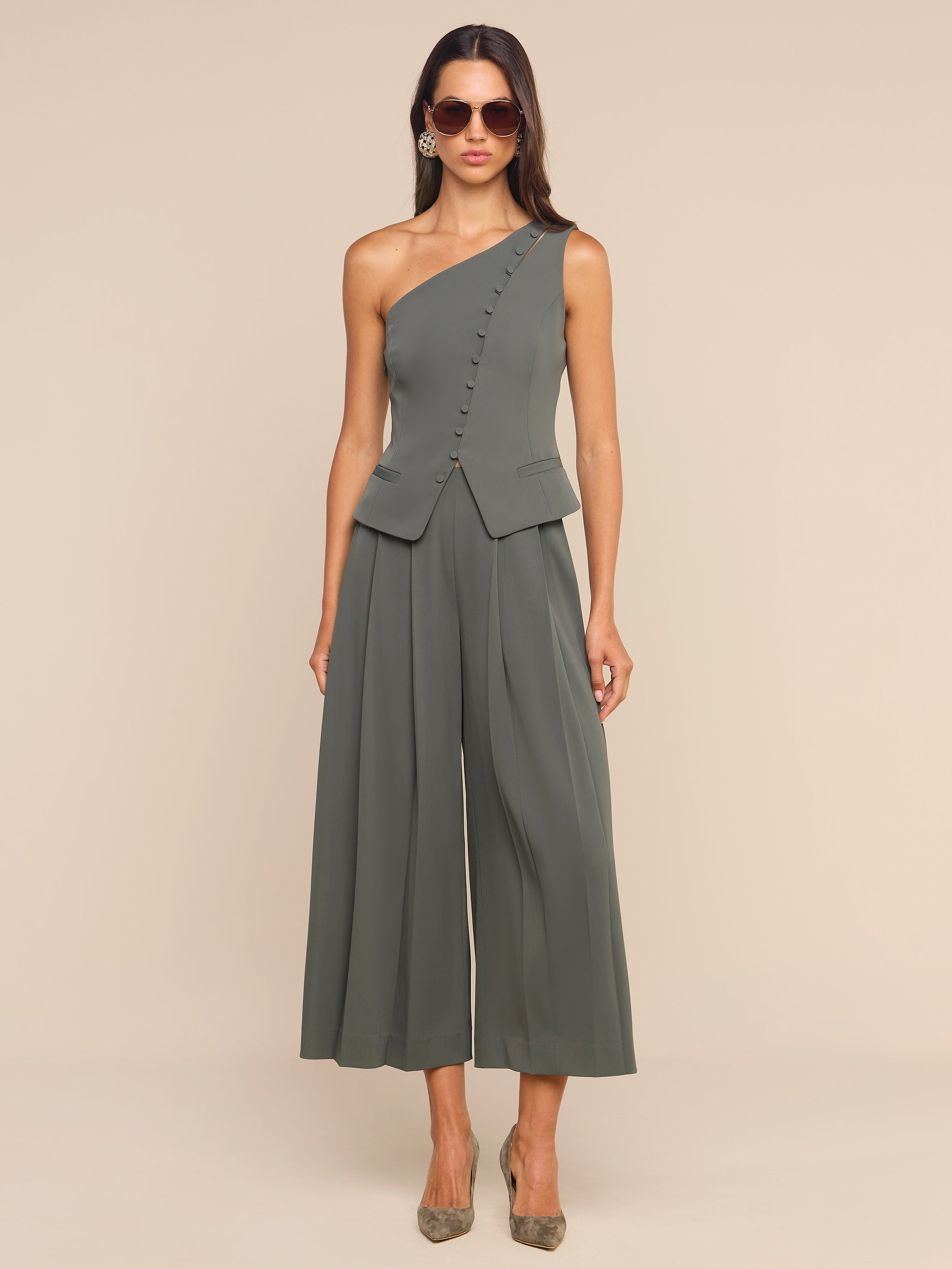 A woman wearing sunglasses and a stylish, olive green, one-shoulder jumpsuit featuring the Novalynn Cropped Pleated Pant with wide legs and buttons, standing against a plain white background.