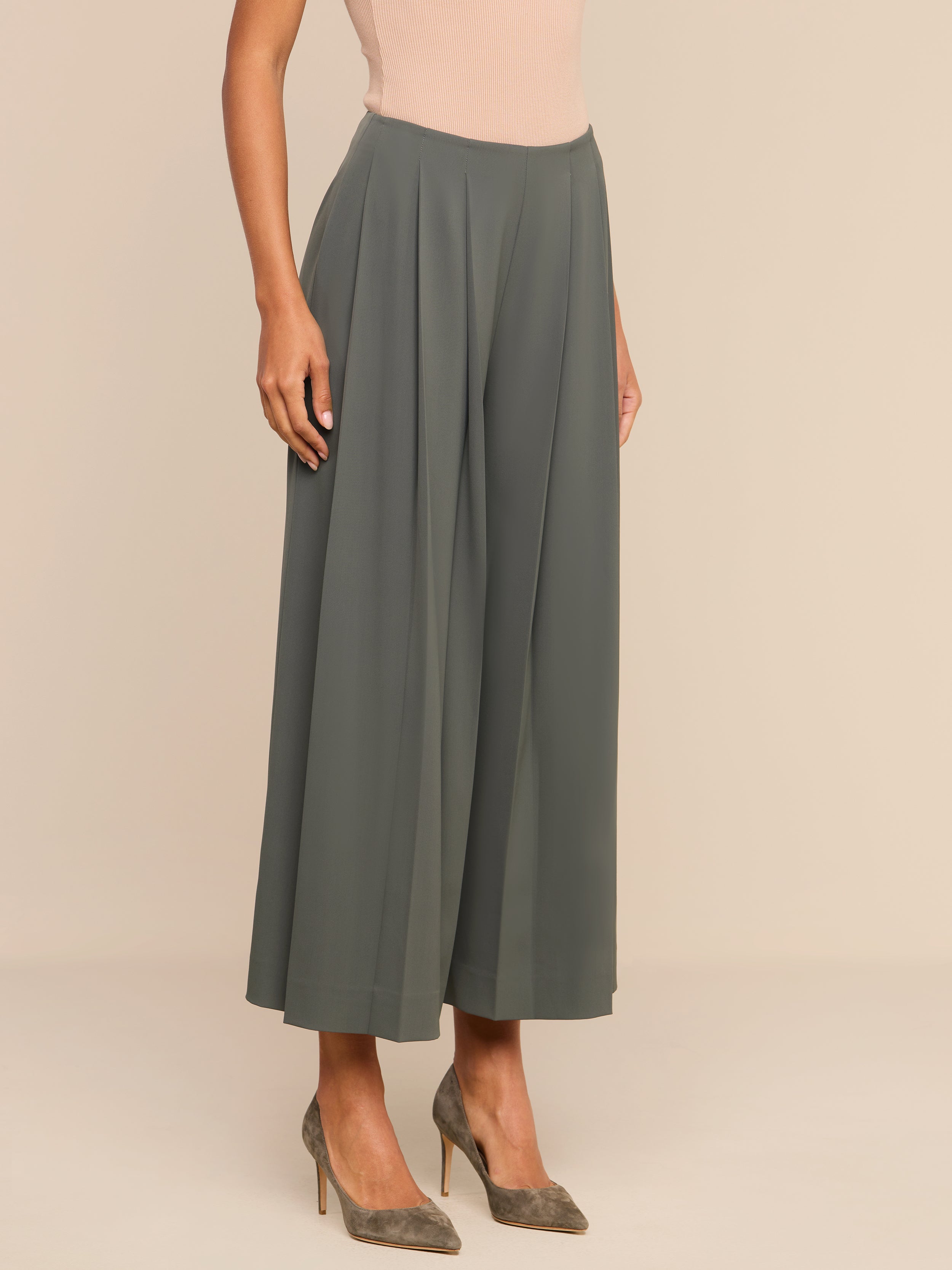 A person wearing loose-fitting, high-rise Novalynn Cropped Pleated Pants in olive green and taupe high heels stands against a plain white background.