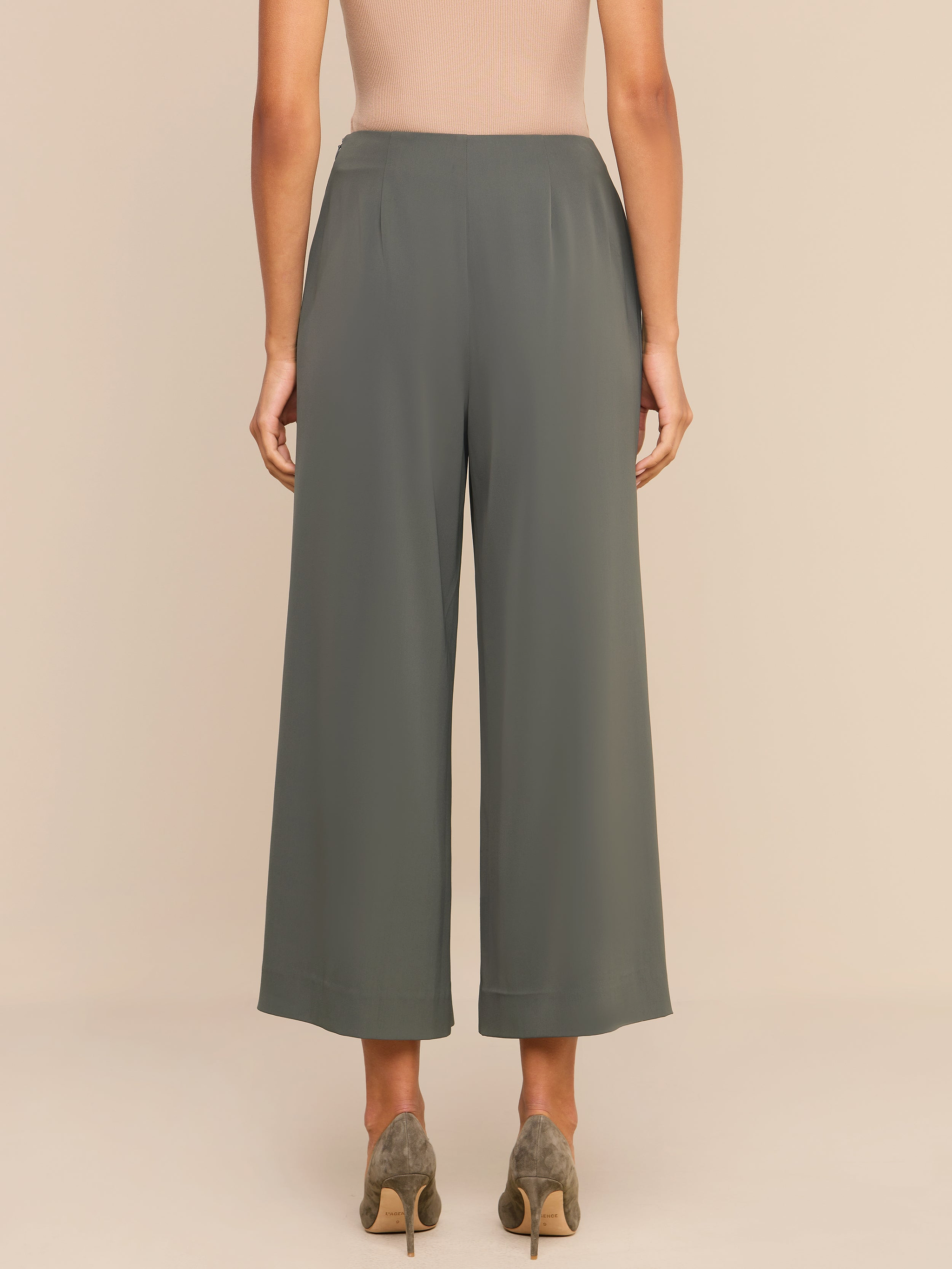 A woman stands facing away, wearing high-rise olive green Novalynn Cropped Pleated Pants with wide legs, a beige sleeveless top, and taupe high heels against a plain white background.