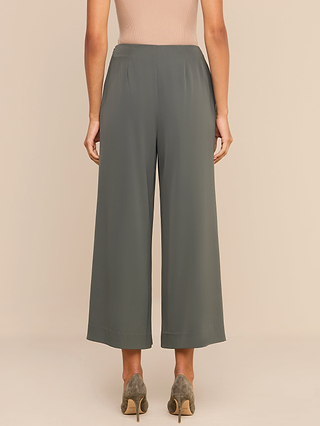 A person stands facing away, wearing the Novalynn Cropped Pleated Pant in olive green with taupe heeled shoes and a beige sleeveless top, set against a plain neutral background.