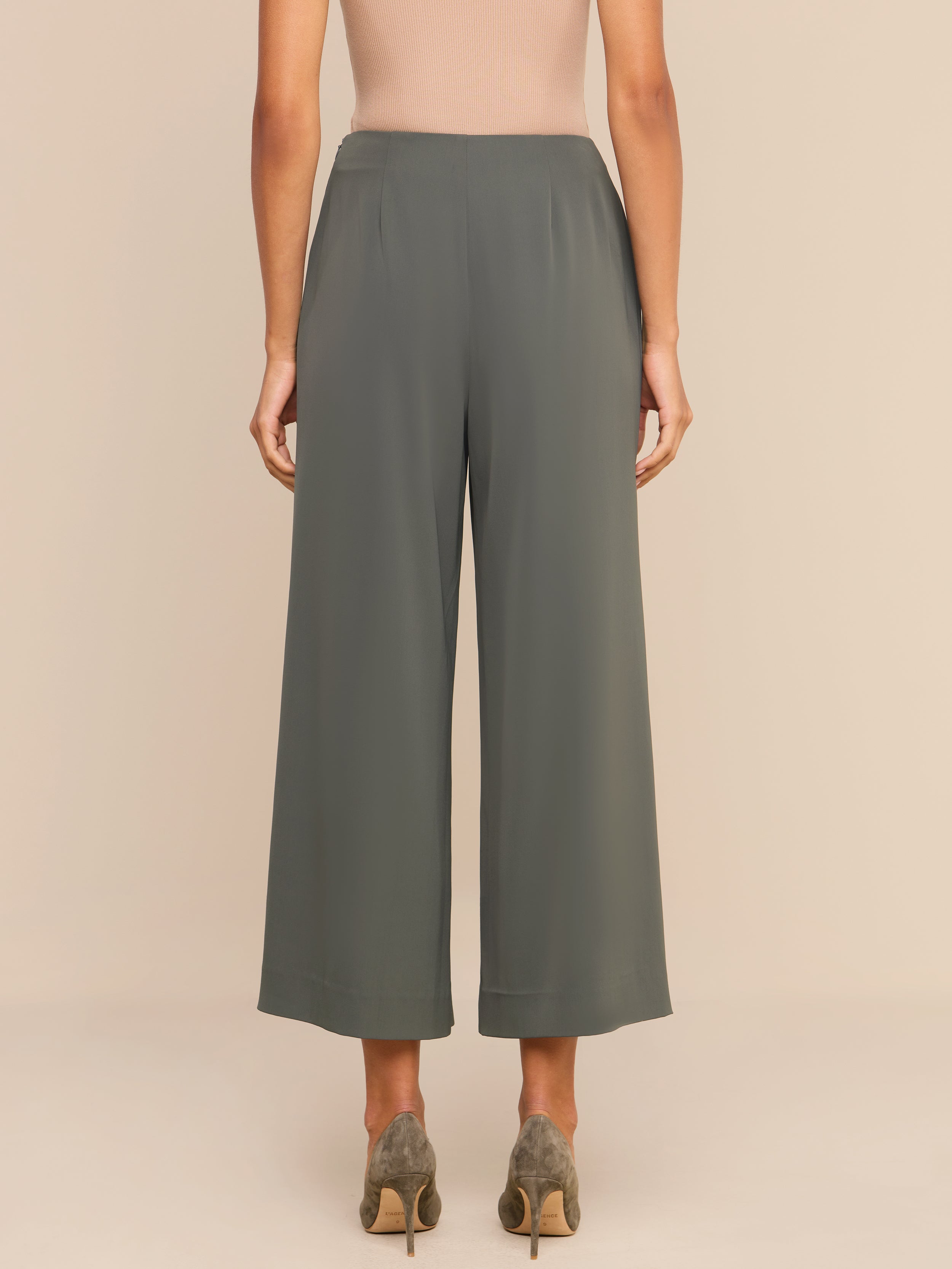 A woman stands facing away, wearing high-rise olive green Novalynn Cropped Pleated Pants with wide legs, a beige sleeveless top, and taupe high heels against a plain white background.