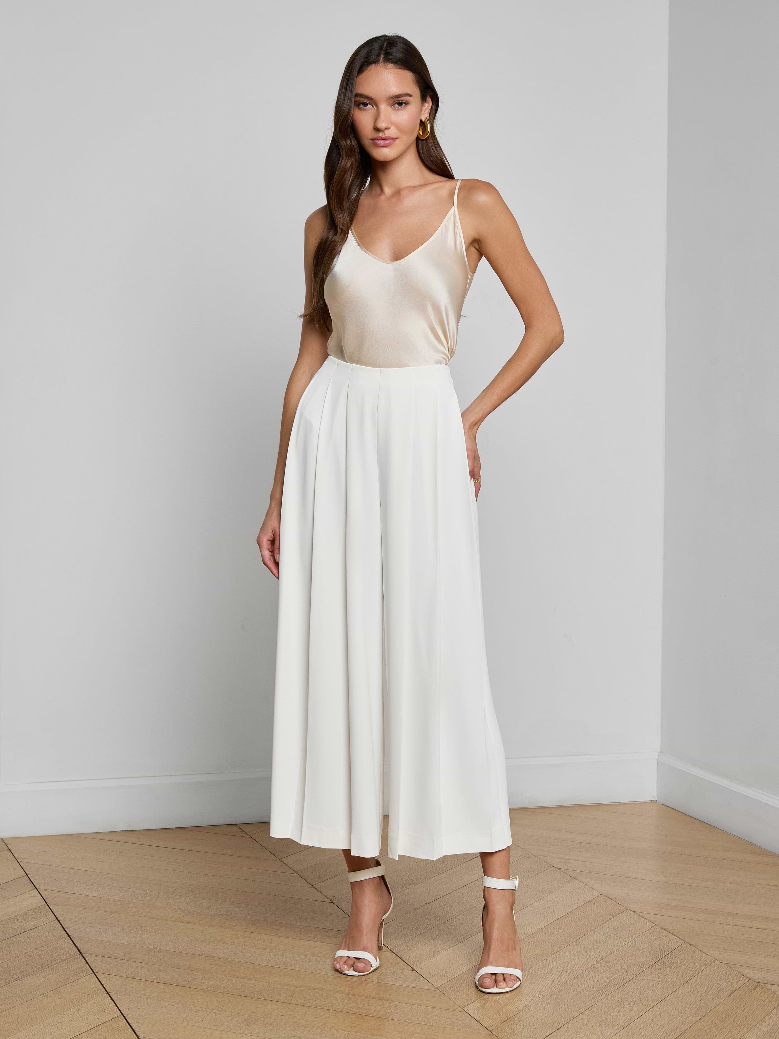 A woman stands against a white background, wearing a sleeveless, light-colored satin top paired with the Novalynn Cropped Pleated Pant and high-heeled sandals.