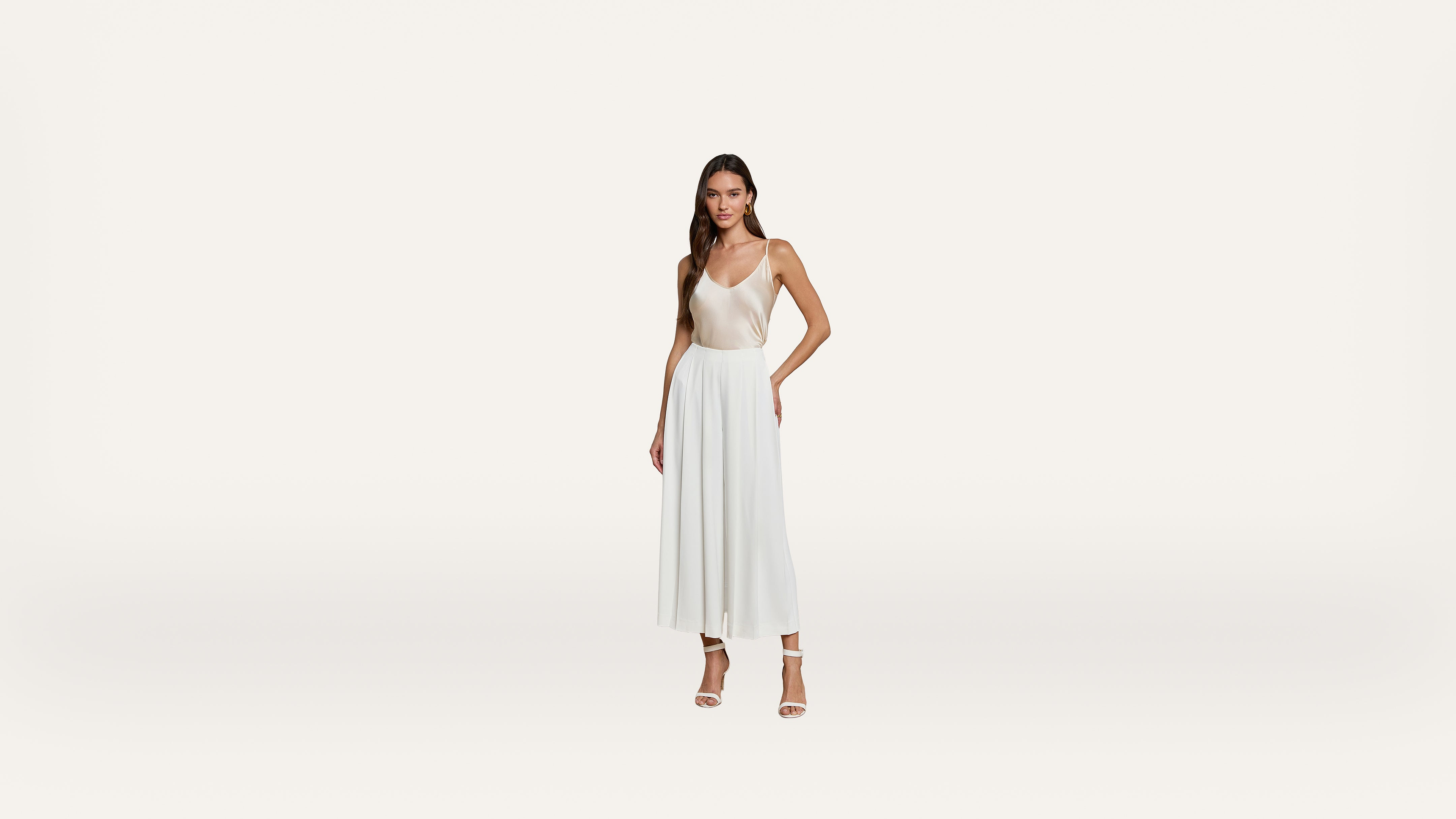 A woman in a white sleeveless dress and heels stands against a plain white background, posing with one hand on her hip.