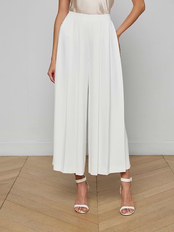 A woman stands against a plain light background wearing the Novalynn Cropped Pleated Pant in white—a high-rise, wide-leg culotte style—paired with a sleeveless beige top and white high-heeled sandals.