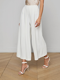 A woman wears the Novalynn Cropped Pleated Pant, a pair of high-rise, wide-leg white trousers, and white open-toe high-heel sandals, with her arms by her sides. The background is plain and light-colored.