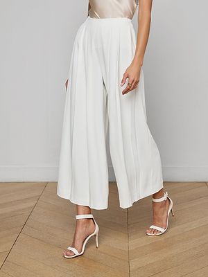 A person wears Novalynn Cropped Pleated Pants, a light top, and white heeled sandals while standing on a wooden floor against a plain light background, with only the lower half of their body visible.