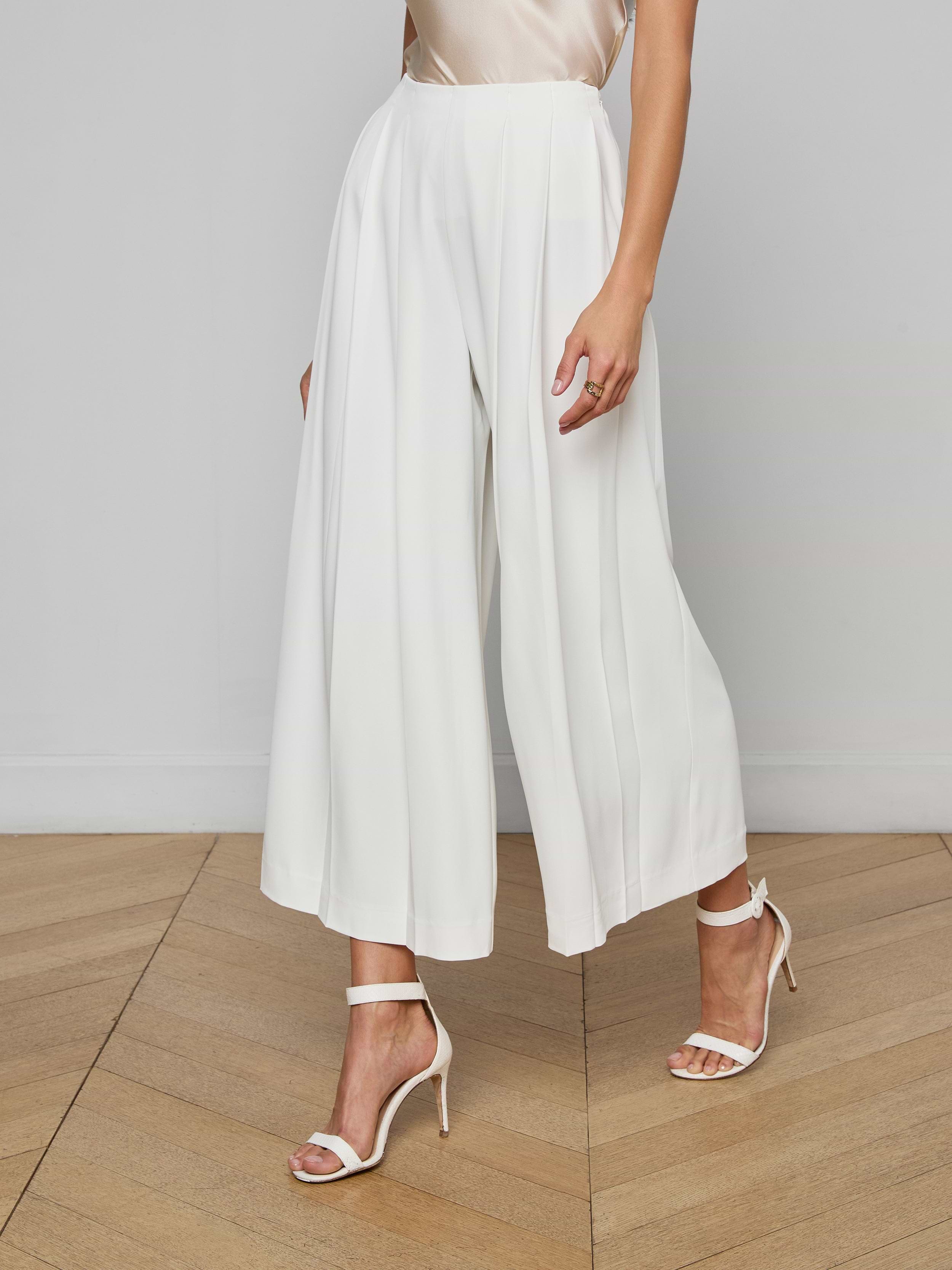 A woman wears the Novalynn Cropped Pleated Pant, a pair of high-rise, wide-leg white trousers, and white open-toe high-heel sandals, with her arms by her sides. The background is plain and light-colored.