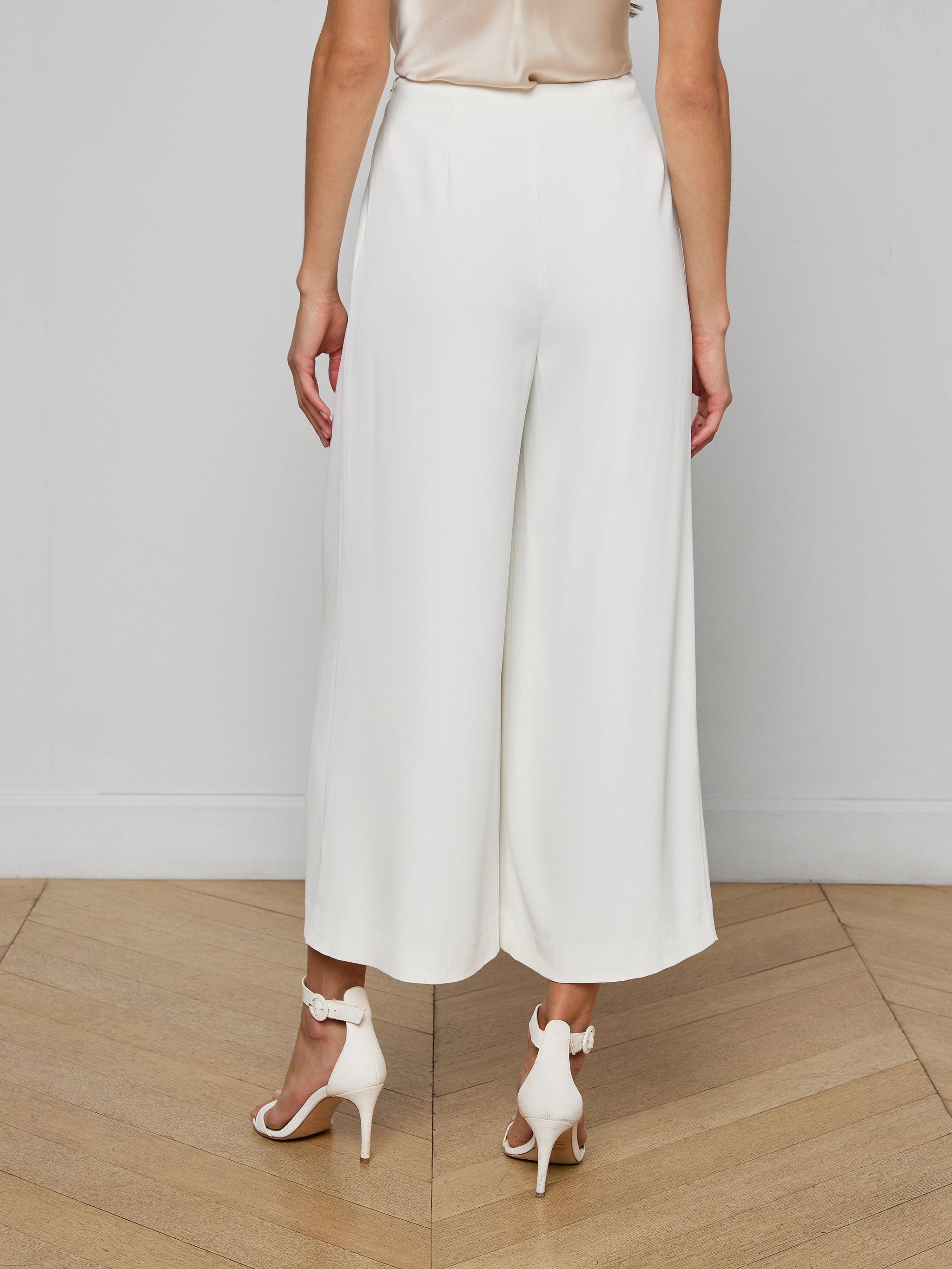 A woman shown from the back against a plain, light background wears the Novalynn Cropped Pleated Pant styled with white high-heeled sandals.