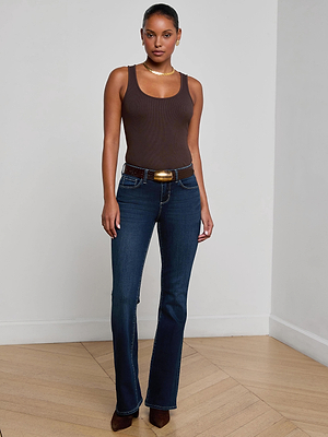 A woman stands indoors wearing L'AGENCE Cara Low-Rise Bootcut Jeans in dark blue super stretch denim, paired with a brown tank top, black belt, brown pointed shoes, and gold jewelry. She poses against a plain white wall on light wood flooring.