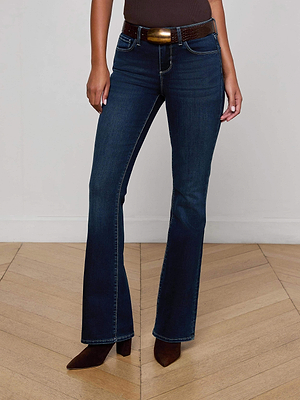A person wearing L'AGENCE Cara Low-Rise Bootcut Jeans in dark blue with a brown belt and pointed-toe shoes stands on a light wooden floor against a plain white wall, shown from shoulders down.