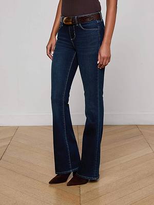 Someone stands on a light wood floor against a plain white wall, wearing L'AGENCE Cara Low-Rise Bootcut Jeans in dark blue super stretch denim, paired with a brown belt, brown pointed-toe shoes, and a dark top.