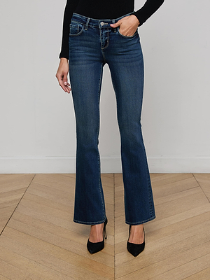 Wearing Cara Low-Rise Bootcut Jeans in dark blue, a black long-sleeve top, and black pointed-toe heels, this individual stands on a wooden floor against a plain white wall.