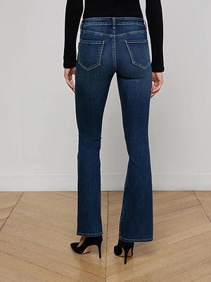 A person stands on a wooden floor facing away from the camera, wearing Cara Low-Rise Bootcut Jeans in dark blue with a black long-sleeve top and black high-heeled shoes, set against a plain white wall.