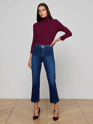 A woman with long dark hair poses with one hand on her hip, wearing a fitted burgundy turtleneck sweater, Noya Cropped Slim Flare Jeans in dark blue wash with front pockets, and burgundy heels against a plain wall.